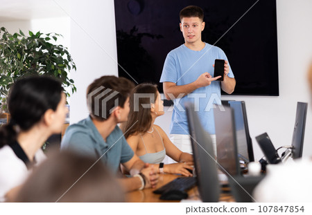 Young male speaker holds mobile phone in hands and explains to listeners of computer courses 107847854