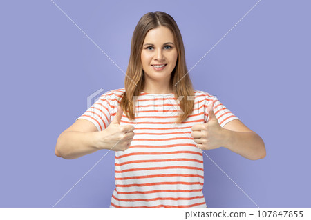 Portrait of blond woman wearing striped T-shirt looking at camera with toothy smile and showing thumbs up, approval sign, satisfied with service. Indoor studio shot isolated on purple background. 107847855