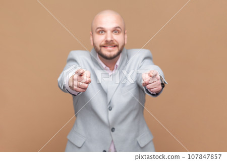 Portrait of cheerful bald bearded man pointing to you, has joyful expression, looking at camera with toothy smile, wearing gray jacket. Indoor studio shot isolated on brown background. Portrait of cheerful bald bearded man pointing to you, has joyful expression, looking at camera with toothy smile, wearing gray jacket. Indoor studio shot isolated on brown background. 107847857