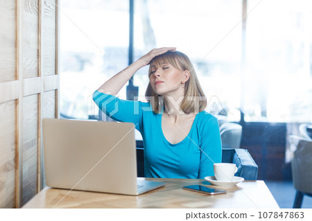 Portrait of sad upset despair young woman with blonde hair in blue shirt working on laptop, making facepalm gesture, has problems with work. Indoor shot in cafe with big window on background. 107847863