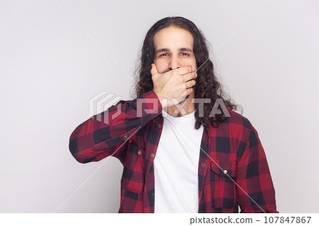 Portrait of bearded man with long curly hair in checkered red shirt tries not tell secret to her friends, covers mouth while gossiping. Indoor studio shot isolated on gray background. 107847867