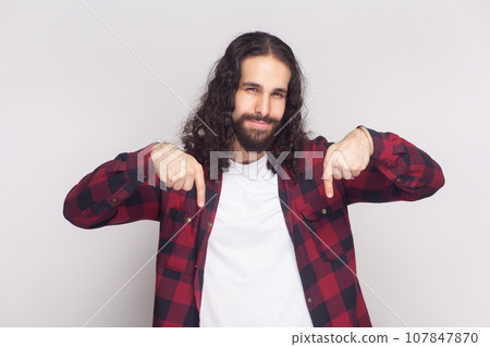 Portrait of bossy anger bearded man with long curly hair in checkered red shirt pointing down and looking arrogant, demanding to do immediately. Indoor studio shot isolated on gray background. 107847870