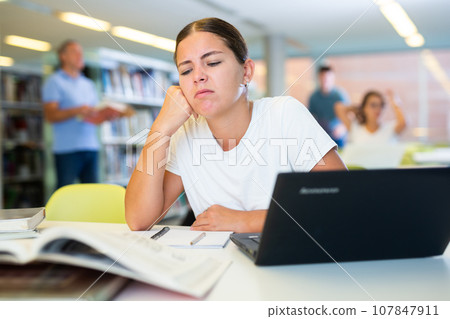 Tired casual female remote worker using laptop and reading books in the library 107847911