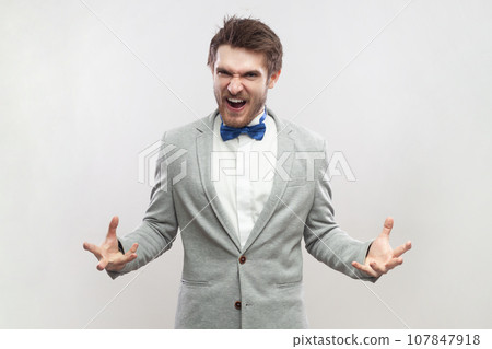 Portrait of angry aggressive handsome bearded man standing with raised arms, screaming, expressing hate and anger, wearing grey suit and blue bow tie. Indoor studio shot isolated on gray background. 107847918