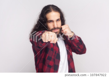 Aggressive bearded man with long curly hair in checkered red shirt keeping his fists ready to fight and defense himself against injustice or violence. Indoor studio shot isolated on gray background. 107847922