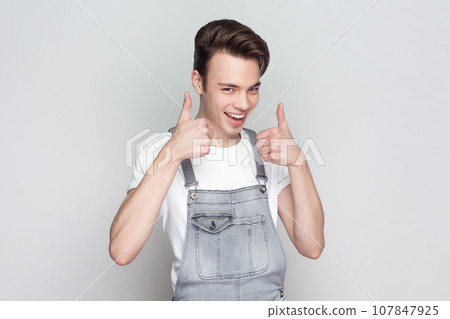 Nice joke. Portrait of young brunette man standing keeps thumb raised, being in good mood, shows his agreement, wearing denim overalls. Indoor studio shot isolated on gray background. Nice joke. Portrait of young brunette man standing keeps thumb raised, being in good mood, shows his agreement, wearing denim overalls. Indoor studio shot isolated on gray background. 107847925