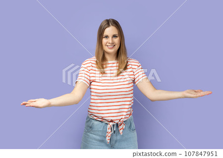 Friendly positive optimistic blond woman wearing striped T-shirt standing with raised arms, looking with toothy smile, offering to take for free. Indoor studio shot isolated on purple background. Friendly positive optimistic blond woman wearing striped T-shirt standing with raised arms, looking with toothy smile, offering to take for free. Indoor studio shot isolated on purple background. 107847951