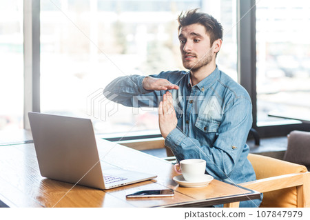 Portrait of handsome bearded young man freelancer in blue jeans shirt working on laptop, showing time out gesture to computer display. Indoor shot near big window, cafe background. 107847979