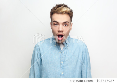 Portrait of amazed surprised man wearing denim shirt looking at camera with big eyes and open mouth, sees something shocked and astonished. Indoor studio shot isolated on gray background. Portrait of amazed surprised man wearing denim shirt looking at camera with big eyes and open mouth, sees something shocked and astonished. Indoor studio shot isolated on gray background. 107847987