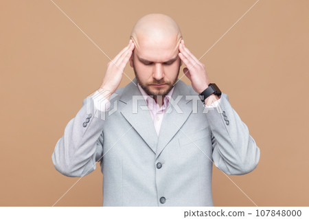 Portrait of sick ill unhealthy bald bearded man standing with hands on head, keeps eyes closed, suffering terrible headache, wearing gray jacket. Indoor studio shot isolated on brown background. Portrait of sick ill unhealthy bald bearded man standing with hands on head, keeps eyes closed, suffering terrible headache, wearing gray jacket. Indoor studio shot isolated on brown background. 107848000