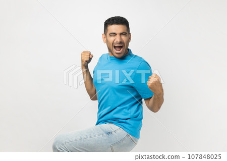 Portrait of unshaven man wearing blue T- shirt standing clenches fists, rejoices his victory and triumph, expresses positiveness, feels like champion. Indoor studio shot isolated on gray background. Portrait of unshaven man wearing blue T- shirt standing clenches fists, rejoices his victory and triumph, expresses positiveness, feels like champion. Indoor studio shot isolated on gray background. 107848025