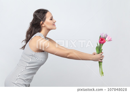 Side view portrait of pretty brunette woman holding out bouquet of tulips, giving it to her mother, congratulating on 8 March, wearing striped dress. Indoor studio shot isolated on gray background. Side view portrait of pretty brunette woman holding out bouquet of tulips, giving it to her mother, congratulating on 8 March, wearing striped dress. Indoor studio shot isolated on gray background. 107848028