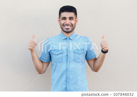 Portrait of extremely happy joyful cheerful man wearing denim shirt looking at camera with toothy smile, showing thumbs up, approved. Indoor studio shot isolated on gray background. Portrait of extremely happy joyful cheerful man wearing denim shirt looking at camera with toothy smile, showing thumbs up, approved. Indoor studio shot isolated on gray background. 107848032