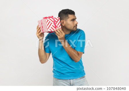 Portrait of curious unshaven man wearing blue T- shirt standing with interested facial expression, shaking present box, thinking what inside. Indoor studio shot isolated on gray background. Portrait of curious unshaven man wearing blue T- shirt standing with interested facial expression, shaking present box, thinking what inside. Indoor studio shot isolated on gray background. 107848040