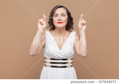 Portrait of pleasant looking hopeful middle aged woman with wavy hair crosses fingers and has great hope for better, wearing white dress. Indoor studio shot isolated on light brown background. 107848073