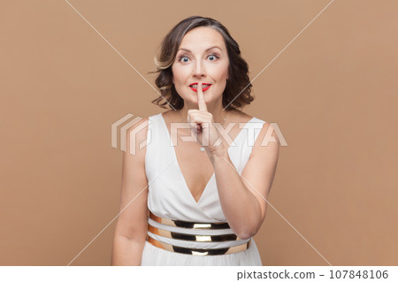 Portrait of woman makes silence or hush gesture, keeps index fingers over lips, tells secret information to someone, wearing white dress. Indoor studio shot isolated on light brown background. 107848106
