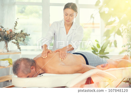 Skilled middle-aged masseuse conducting back massage for man client 107848120