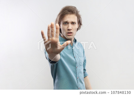 Portrait of young man making stop gesture showing palm of hand, conflict prohibition warning about danger, stop bullying, wearing blue shirt. Indoor studio shot isolated on gray background. 107848130