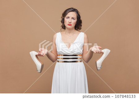 Portrait of sad upset middle aged woman with wavy hair holding elegant shoes on high heels, feels pain on feet, wearing white dress. Indoor studio shot isolated on light brown background. 107848133