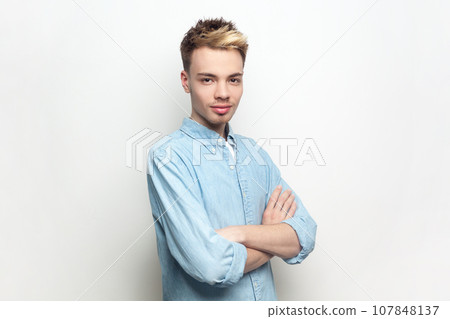 Portrait of self-confident attractive handsome man wearing denim shirt standing with crossed arms, looking at camera, expressing positive emotions. Indoor studio shot isolated on gray background. 107848137