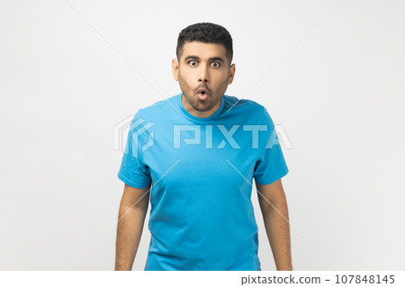Portrait of amazed shocked surprised unshaven man wearing blue T- shirt standing looking at camera with big eyes, sees something astonished. Indoor studio shot isolated on gray background. Portrait of amazed shocked surprised unshaven man wearing blue T- shirt standing looking at camera with big eyes, sees something astonished. Indoor studio shot isolated on gray background. 107848145