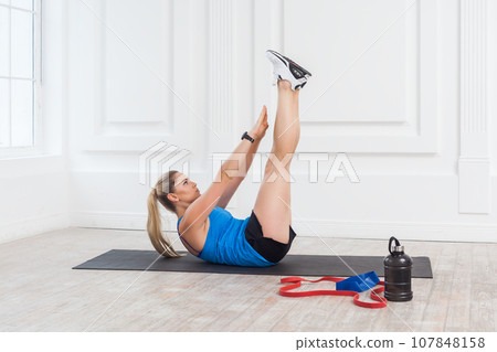 Sport and healthy concept. Side view portrait of strong sporty beautiful blonde woman in black shorts and blue top working in gym, doing exercise for perfect press. Indoor studio shot. Sport and healthy concept. Side view portrait of strong sporty beautiful blonde woman in black shorts and blue top working in gym, doing exercise for perfect press. Indoor studio shot. 107848158