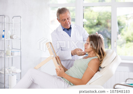 Woman looking at mirror while elderly cosmetologist showing results of facial procedure Woman looking at mirror while elderly cosmetologist showing results of facial procedure 107848183