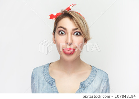 Portrait of funny crazy foolish blonde woman wearing blue denim shirt and red headband standing sticking tongue out, looking at camera. Indoor studio shot isolated on gray background. Portrait of funny crazy foolish blonde woman wearing blue denim shirt and red headband standing sticking tongue out, looking at camera. Indoor studio shot isolated on gray background. 107848194