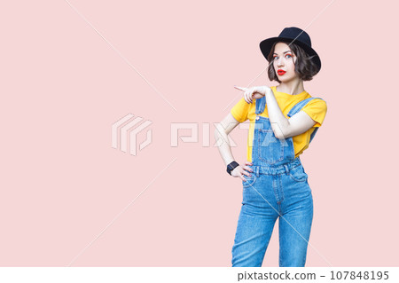 Portrait of surprised amazed hipster woman in blue denim overalls, yellow T-shirt and black hat, pointing at copy space for advertisement. Indoor studio shot isolated on light pink background. 107848195