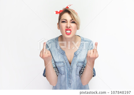 Portrait of angry aggressive rude blonde woman wearing blue denim shirt and red headband standing showing fuck sign, arguing with somebody. Indoor studio shot isolated on gray background. 107848197