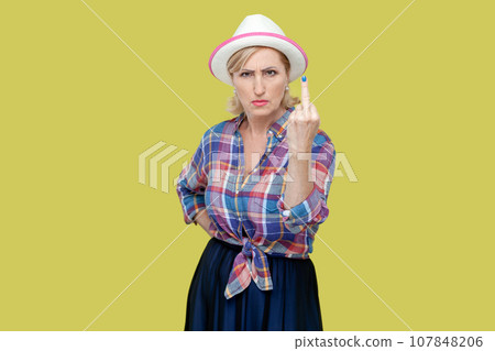 Portrait of mature woman wearing checkered shirt and hat shows middle finger impolite gesture disrespects someone looks rude at camera. Indoor studio shot isolated on yellow background. 107848206