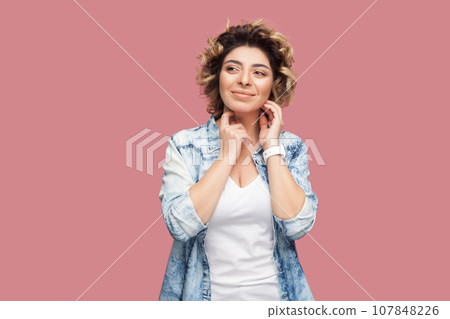 Portrait of smiling beautiful woman with curly hairstyle wearing blue shirt standing looking away, smiling, dreaming about something pleasant. Indoor studio shot isolated on pink background. Portrait of smiling beautiful woman with curly hairstyle wearing blue shirt standing looking away, smiling, dreaming about something pleasant. Indoor studio shot isolated on pink background. 107848226