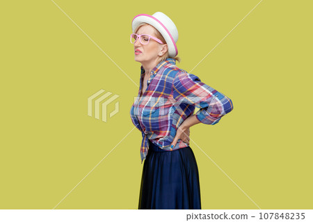 Senior woman wearing checkered shirt, hat and eyeglasses suffers from radiculitis, keeps hand on waist, problems with health, has pain in spine. Indoor studio shot isolated on yellow background. 107848235