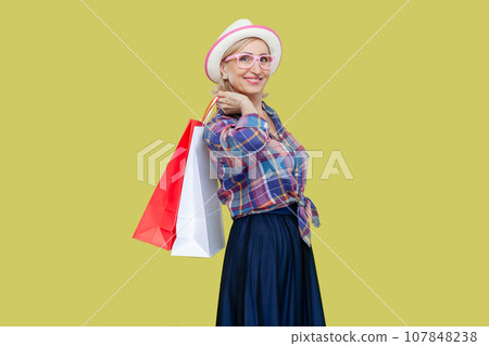 Side view portrait of joyful cheerful mature woman wearing checkered shirt, hat and eyeglasses holding shopping bag, enjoying. Indoor studio shot isolated on yellow background. 107848238