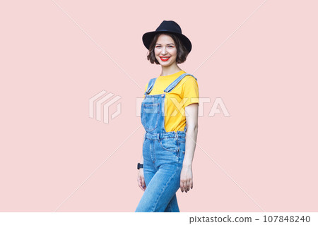 Portrait of cheerful positive hipster woman in blue denim overalls, yellow T-shirt and black hat, looking at camera with toothy smile. Indoor studio shot isolated on light pink background. Portrait of cheerful positive hipster woman in blue denim overalls, yellow T-shirt and black hat, looking at camera with toothy smile. Indoor studio shot isolated on light pink background. 107848240