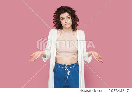 Portrait of puzzled uncertain woman with curly hairstyle wearing casual style outfit spreads hands aside, doesn't know answer. Indoor studio shot isolated on pink background. Portrait of puzzled uncertain woman with curly hairstyle wearing casual style outfit spreads hands aside, doesn't know answer. Indoor studio shot isolated on pink background. 107848247