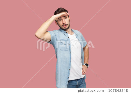 Bearded man in blue casual style shirt standing keeps palm near forehead, looks with concentrated expression into distance, sees something aside. Indoor studio shot isolated on pink background. 107848248