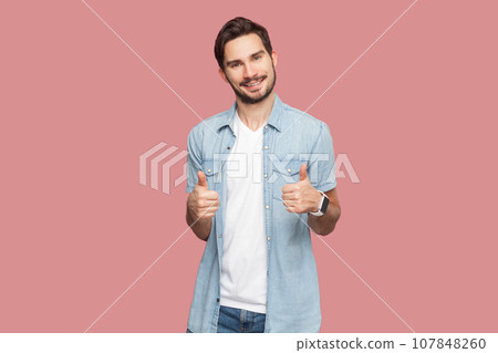 Man in blue shirt gesturing thumbs-up, telling you that you are doing a good job, looking and smiling at the camera with cheerful expression. Indoor studio shot isolated on pink background. 107848260