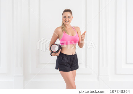 Portrait of sporty young beautiful woman with perfect body wearing pink top and black shorts standing, holding foam roll and showing thumb up, looking at camera. Indoor studio shot. Portrait of sporty young beautiful woman with perfect body wearing pink top and black shorts standing, holding foam roll and showing thumb up, looking at camera. Indoor studio shot. 107848264