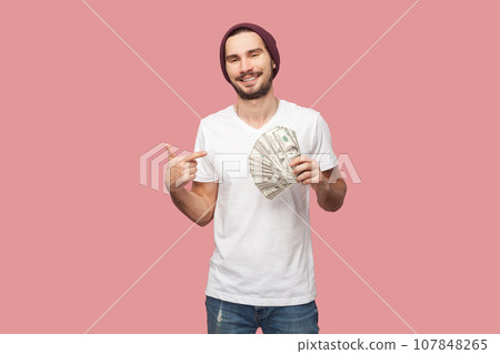 Portrait of satisfied delighted bearded man in white T-shirt and beany hat standing pointing at dollar banknotes, looking smiling at camera. Indoor studio shot isolated on pink background. 107848265