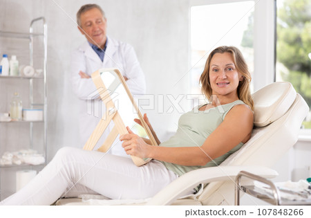 Satisfied middle-aged woman holding mirror lying on clinical chair 107848266
