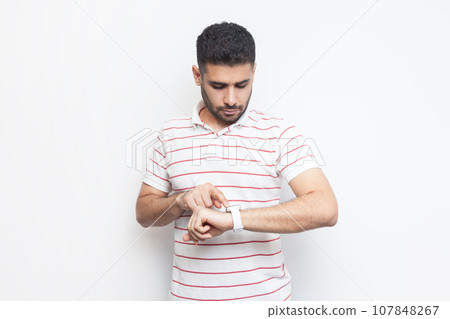 Portrait of concentrated attractive handsome bearded man wearing striped t-shirt standing using his smart watch, uses application. Indoor studio shot isolated on gray background. Portrait of concentrated attractive handsome bearded man wearing striped t-shirt standing using his smart watch, uses application. Indoor studio shot isolated on gray background. 107848267