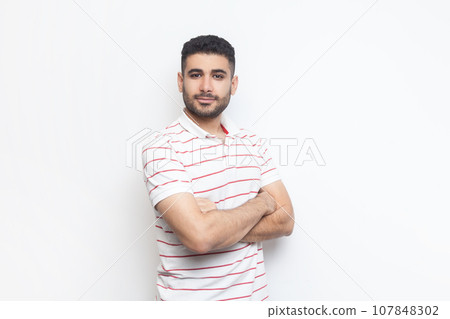 Portrait of delighted positive confident attractive bearded man wearing striped t-shirt standing with crossed hands, looking at camera. Indoor studio shot isolated on gray background. 107848302