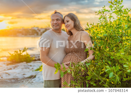 A happy, mature couple over 40, enjoying a leisurely walk on the waterfront On the Sunset, their joy evident as they embrace the journey of pregnancy later in life A happy, mature couple over 40, enjoying a leisurely walk on the waterfront On the Sunset, their joy evident as they embrace the journey of pregnancy later in life 107848314