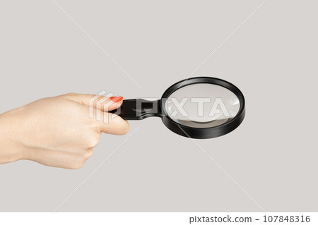 Closeup of woman hand showing loupe, magnificing glass, equipment for inspecting and zooming. Indoor studio shot isolated on gray background. Closeup of woman hand showing loupe, magnificing glass, equipment for inspecting and zooming. Indoor studio shot isolated on gray background. 107848316