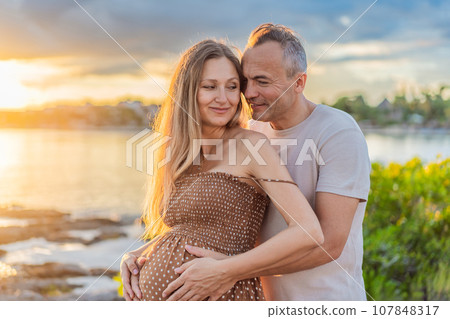 A happy, mature couple over 40, enjoying a leisurely walk on the waterfront On the Sunset, their joy evident as they embrace the journey of pregnancy later in life A happy, mature couple over 40, enjoying a leisurely walk on the waterfront On the Sunset, their joy evident as they embrace the journey of pregnancy later in life 107848317