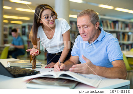 Happy casual Latin woman in glasses talking with older man reading in the library Happy casual Latin woman in glasses talking with older man reading in the library 107848318