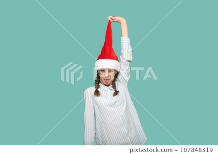 Portrait of happy funny teenager girl with braids wearing striped shirt, looking at camera with tongue out, pulling up her Santa Claus hat. Indoor studio shot isolated on green background. Portrait of happy funny teenager girl with braids wearing striped shirt, looking at camera with tongue out, pulling up her Santa Claus hat. Indoor studio shot isolated on green background. 107848319
