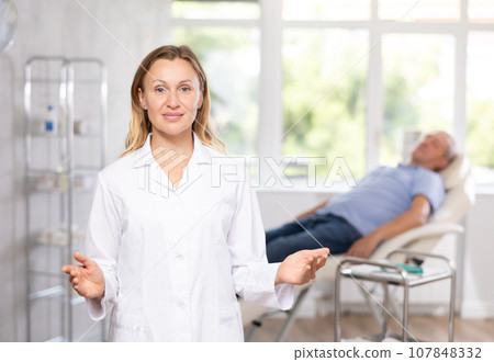 Middle-aged woman doctor posing in doctor's cabinet with patient lying on clinical chair Middle-aged woman doctor posing in doctor's cabinet with patient lying on clinical chair 107848332