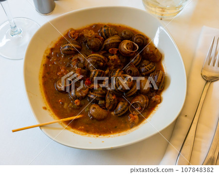 Flavored snails in shells in the tomato sauce in the white ceramic plate on the tablecloth presented in a cafe 107848382